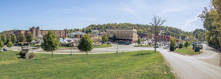 Small-town downtown and park viewed from a grassy hill, with shops, crowds, parked cars, and wooded hills.