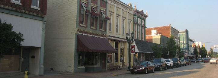 Historic downtown street lined with colorful storefronts, parked cars, trees, and decorative lampposts.