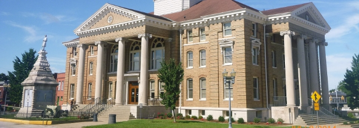 Historic brick courthouse with grand columns, broad steps, landscaped grounds, and a nearby monument.