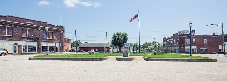 Town square with an American flag, landscaped green space, and historic brick buildings under a clear blue sky.