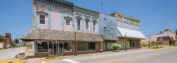 Historic downtown storefronts with restored brick buildings, large display windows, and a wide main street under a clear blue sky.
