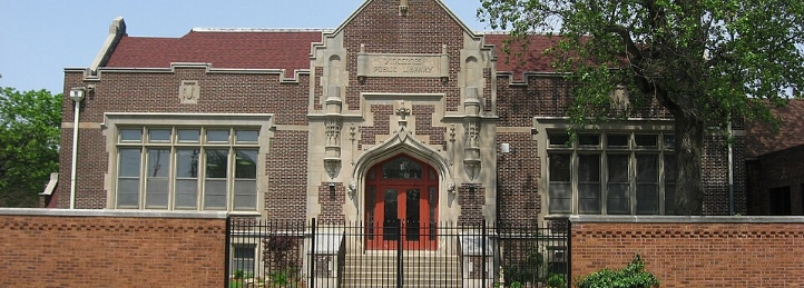 Historic brick school building with arched red entrance, large windows, iron fence, and mature trees.