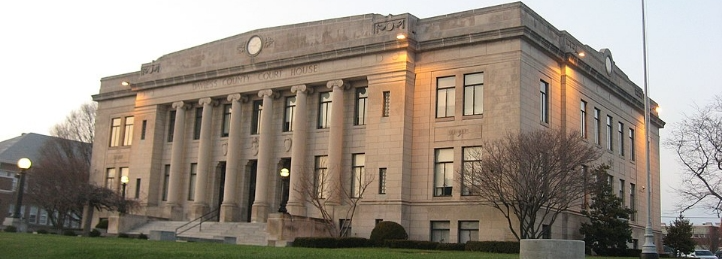 Stone county courthouse with tall columns, broad steps, landscaped lawn, and illuminated exterior.